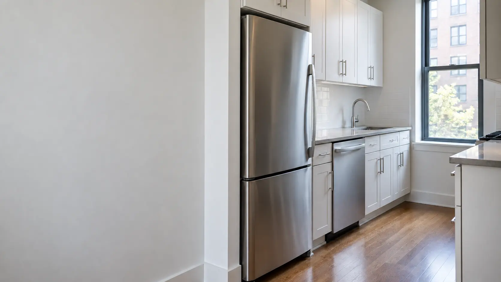 A New York apartment galley kitchen with a slim refrigerator and a dishwasher, seen from across the room.
